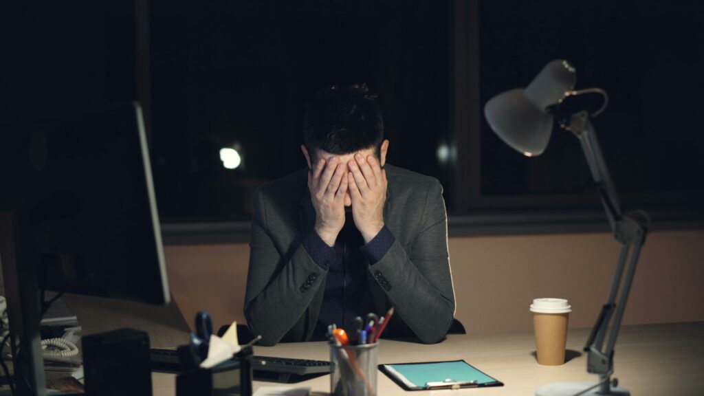 A stressed man sitting at a desk at night with his face in his hands, surrounded by office equipment and paperwork.