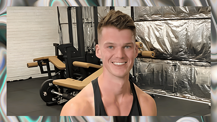 Young man in black tank top smiling in a home gym with weight equipment and foil-wrapped machinery in the background.
